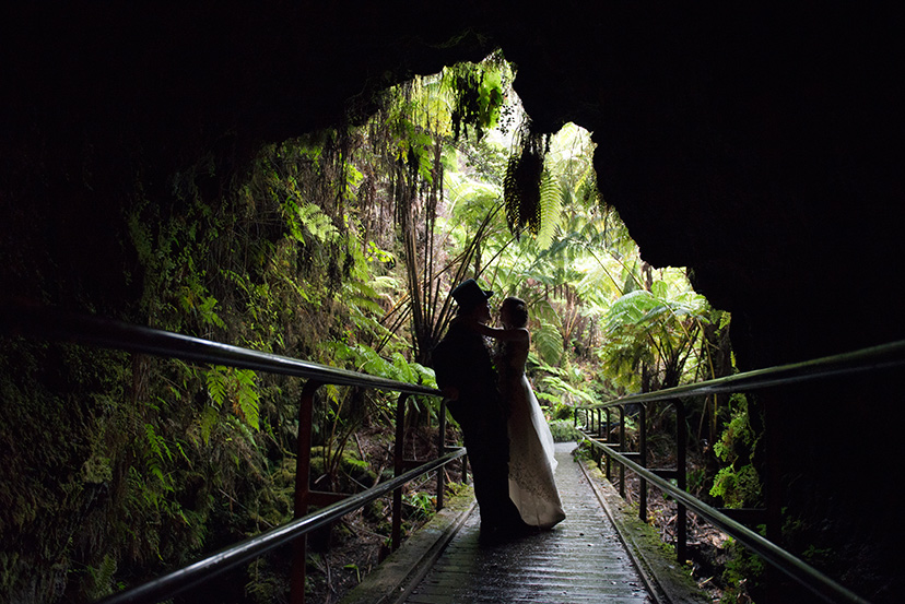 Bride and groom silhouetted in the mouth of a Big Island lava tube with tree ferns in the light beyond