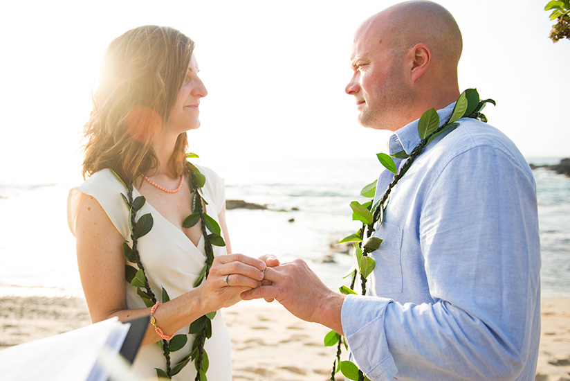 Bride and groom exchanging rings on a Kailua-Kona beach, both wearing green maile leis